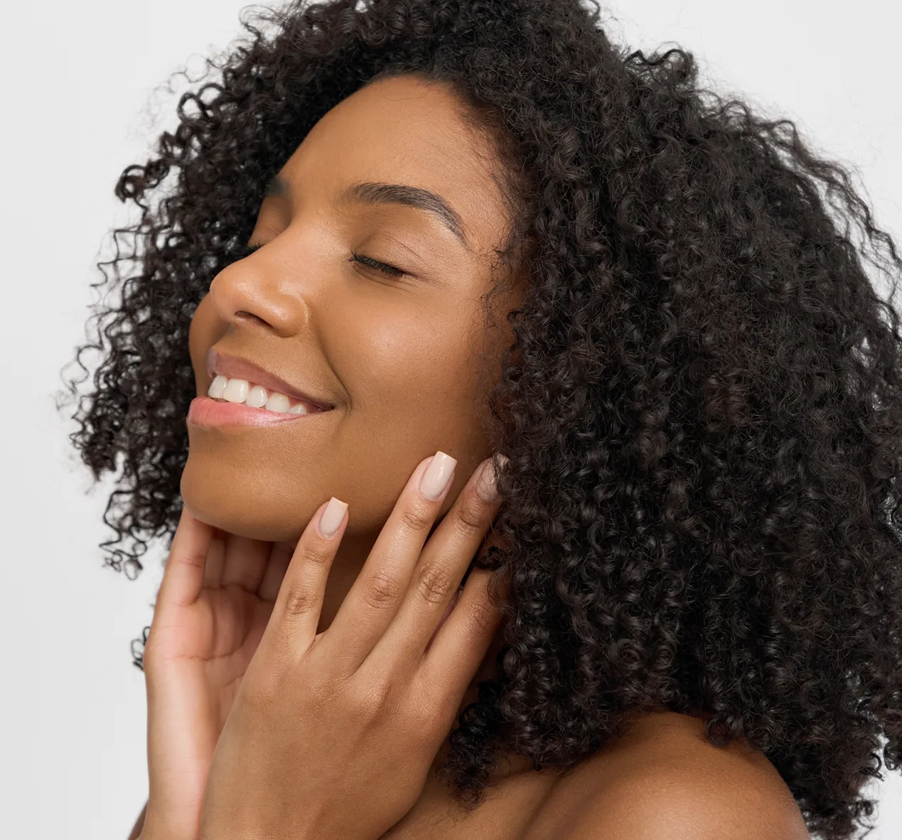 Woman with clear skin and curly dark hair touching her face and smiling